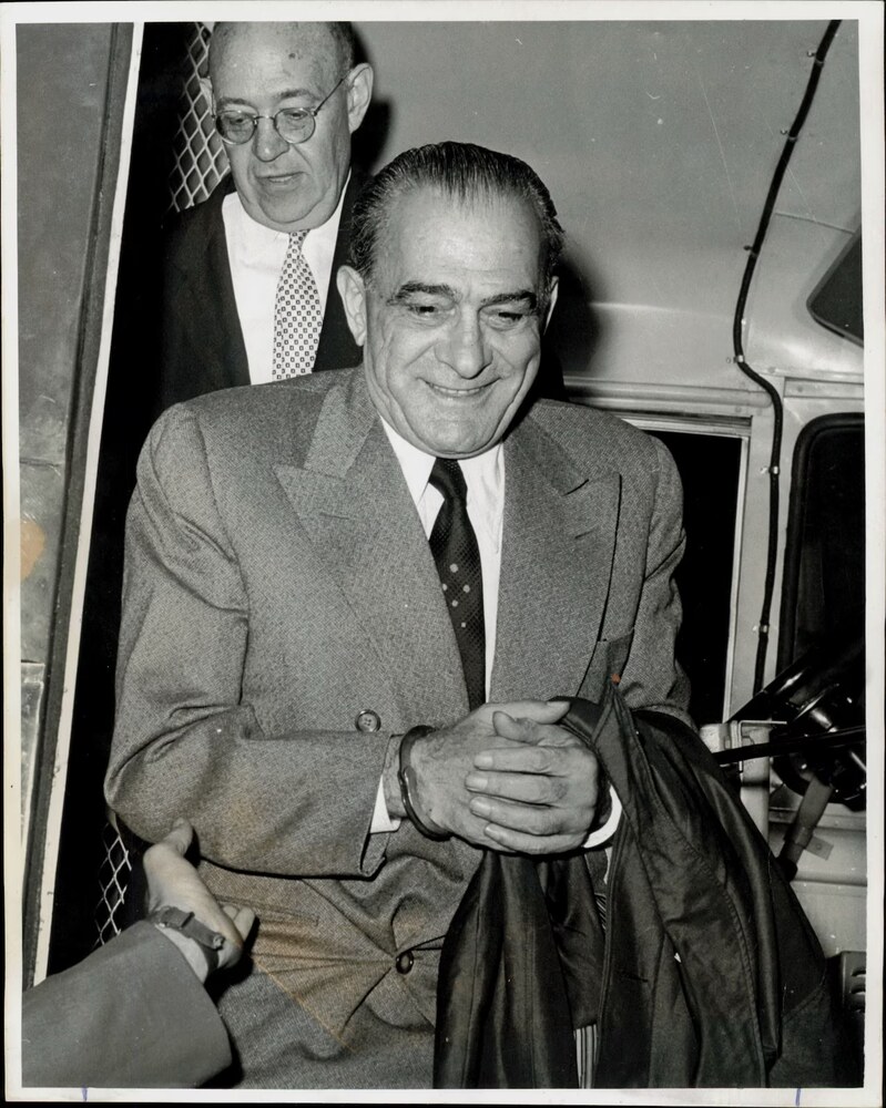 Black-and-white press photograph of mobster Vito Genovese in handcuffs, escorted by a detective