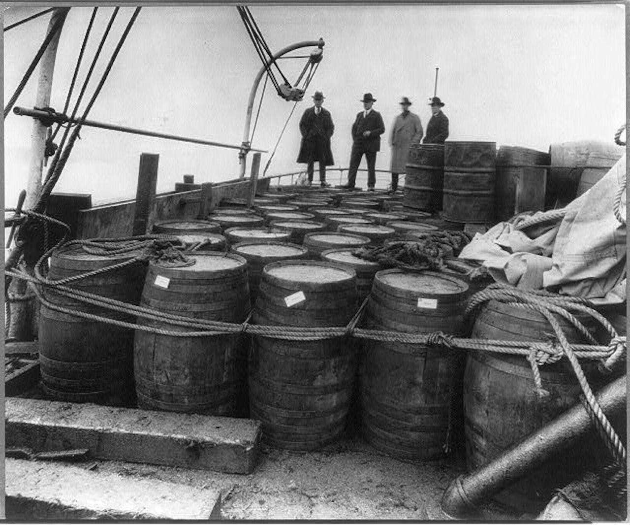 Black-and-white Prohibition-era photograph of men in hats and coats standing among barrels of liquor lashed to a boat deck